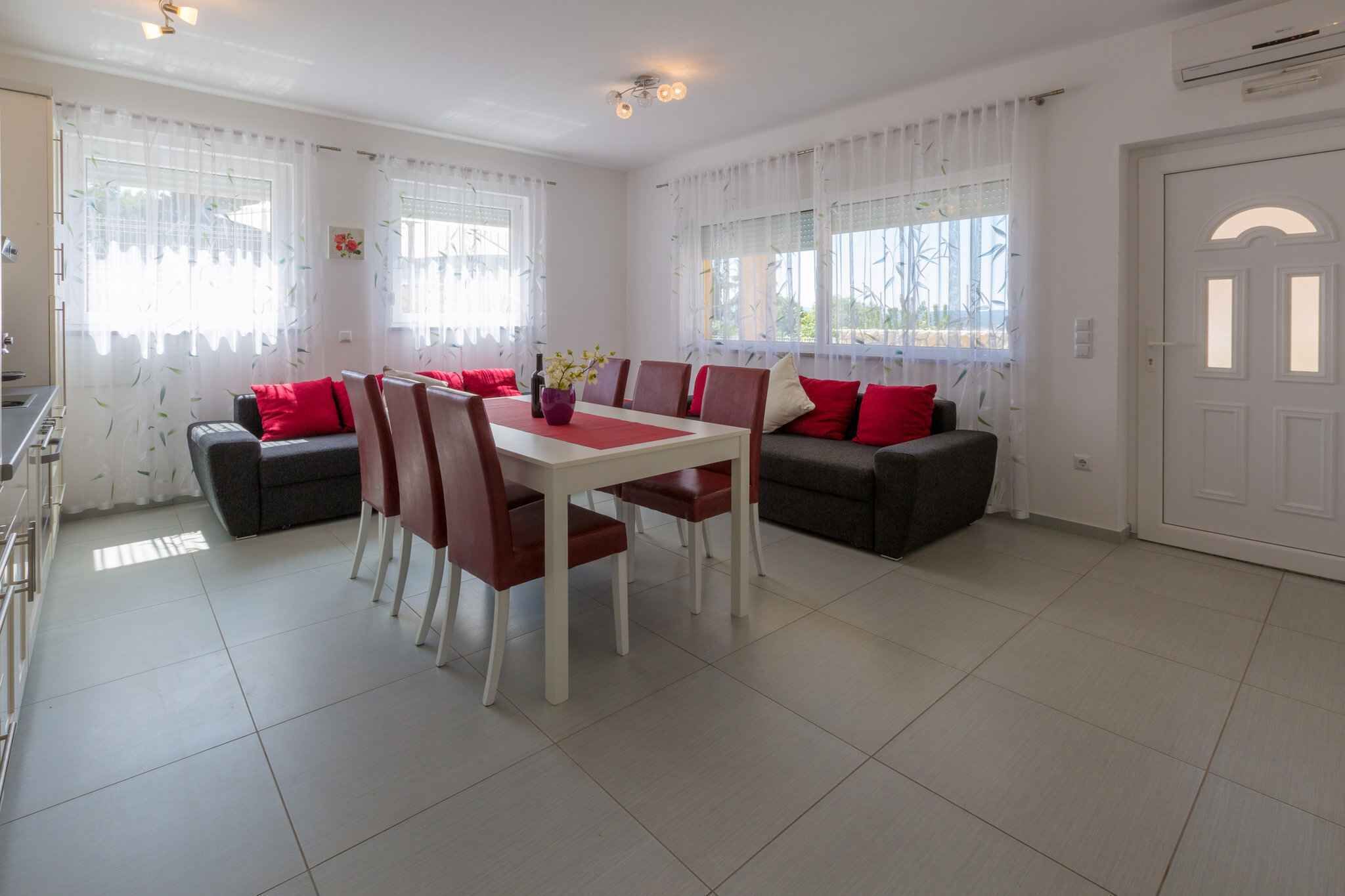 Un comedor con una mesa blanca y sillas rojas, rodeado de sofás con cojines rojos y ventanas amplias con cortinas traslúcidas.