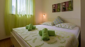 Ein Hotelzimmer mit zwei Betten, grünen Handtüchern, einem Fenster mit transparenten Vorhängen und Wandbildern.