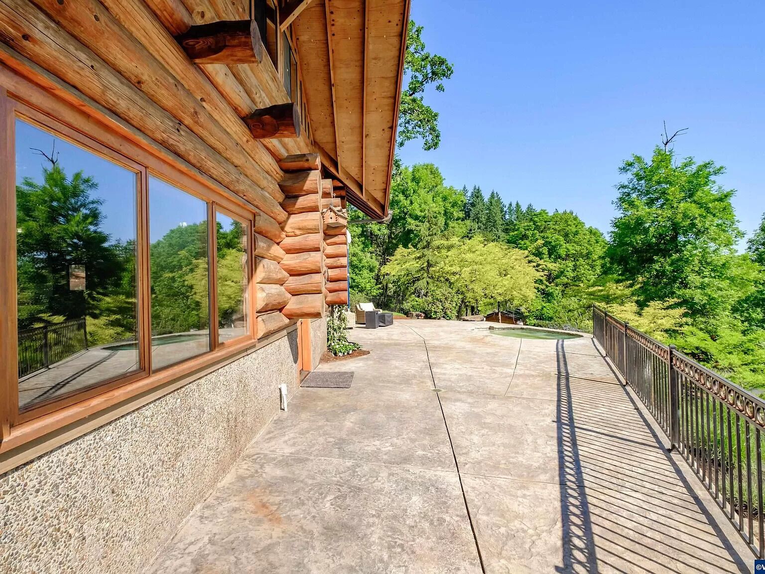 Uma cabana de toras com grandes janelas, um deck de concreto e um guarda-corpo de metal com vista para uma área arborizada.