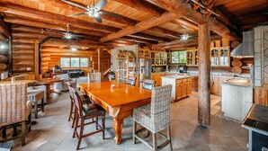Un intérieur spacieux de chalet en bois, avec une grande table à manger, des chaises en osier, un coin cuisine et un ventilateur de plafond.