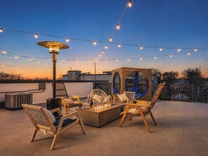 Un área de estar al aire libre con sillas, una chimenea, luces colgantes y vistas del horizonte de la ciudad.