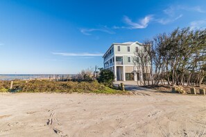 Une maison à deux étages située en bord de mer, dotée d’un balcon, avec une plage de sable en premier plan et un ciel bleu dégagé.