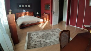 Een slaapkamer met een bed, een houten dressoir, een nachtkastje, een bureau, een stoel, een plafondventilator en een deur die naar een andere kamer leidt.