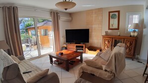 Een woonkamer met een bank, fauteuil en salontafel. Er is een televisie, een kast en een raam met gordijnen.