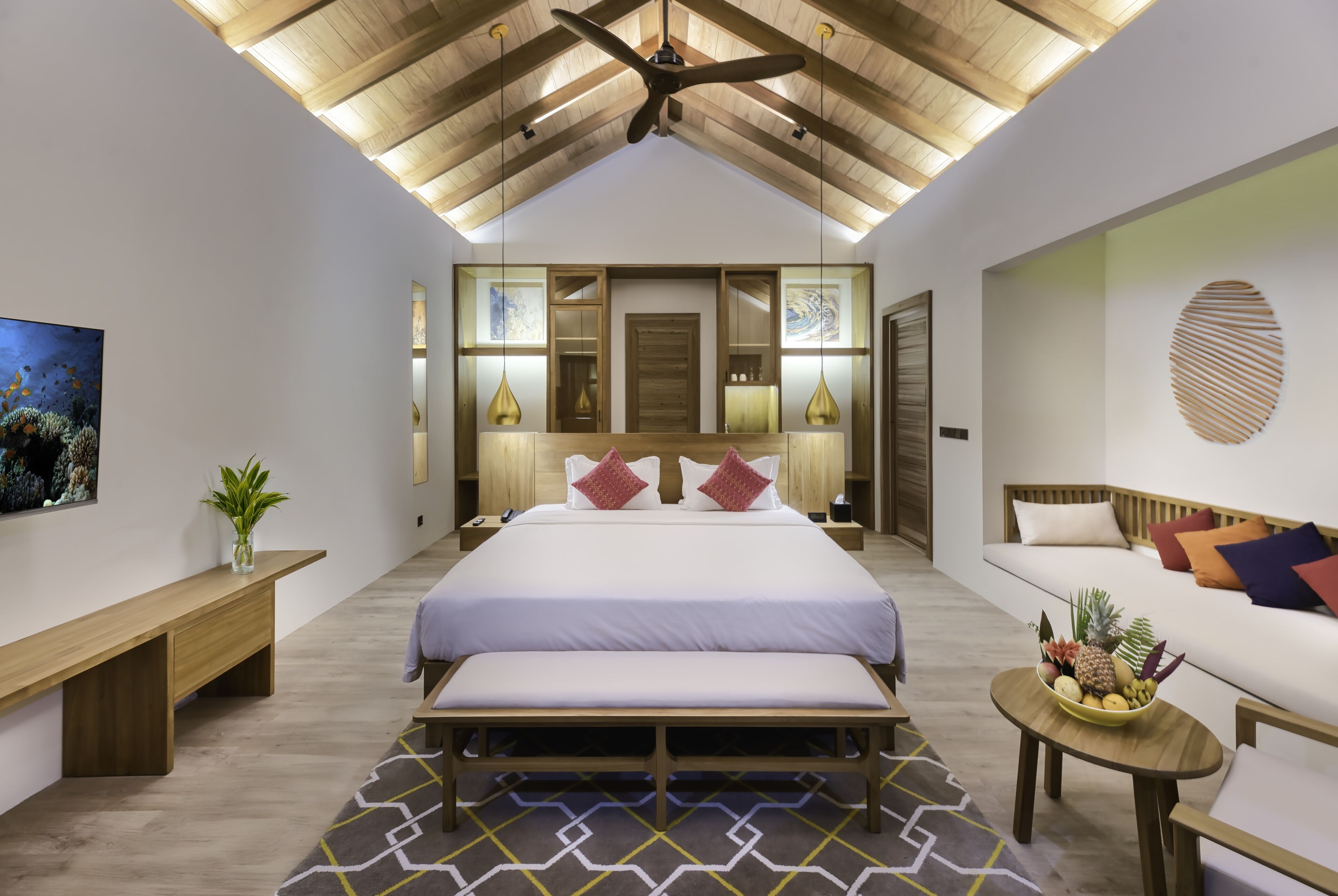 A modern bedroom with a large bed, a bench, a small table with fruit, and a ceiling fan.