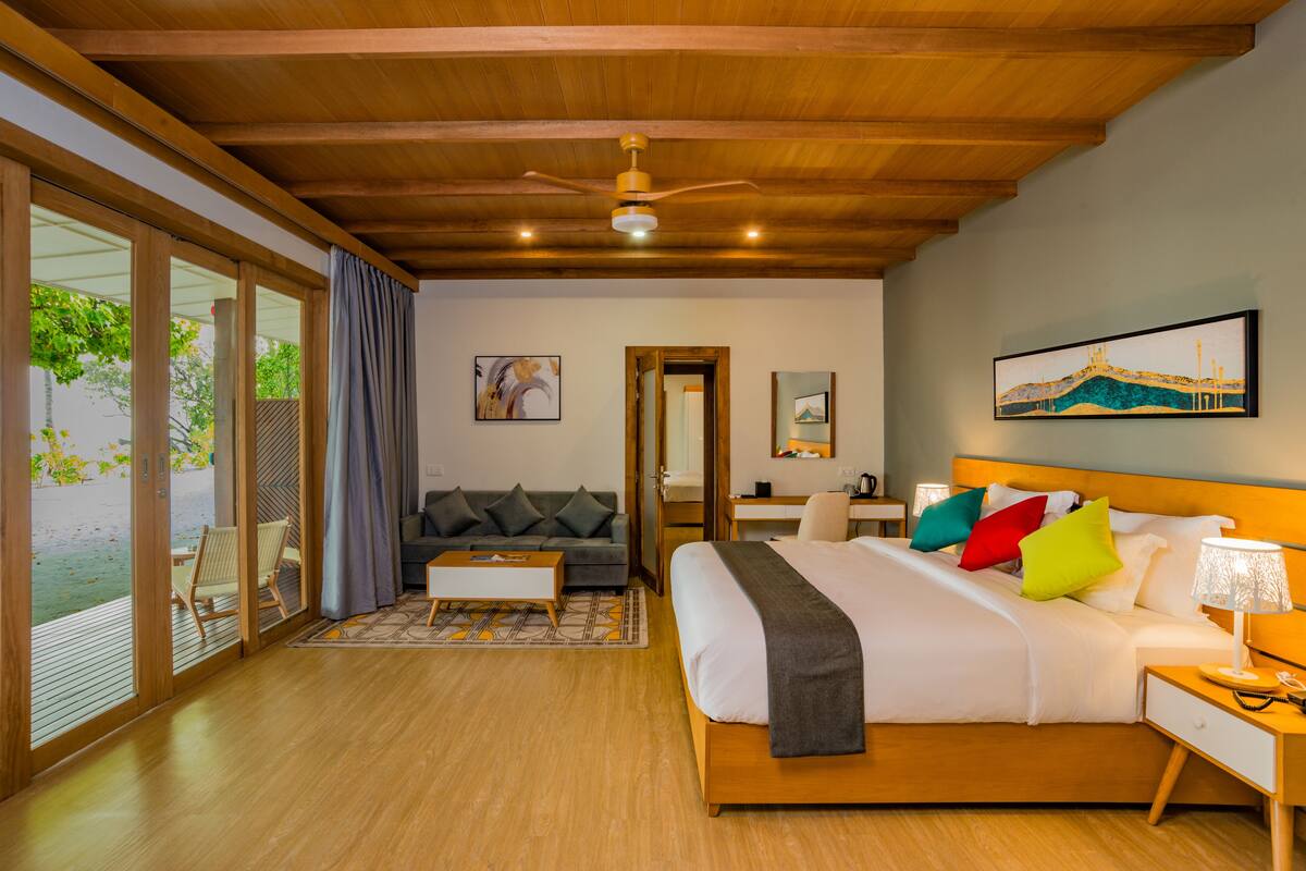 A modern bedroom with a large bed, a desk, and a sofa. There is a sliding glass door leading to a balcony.