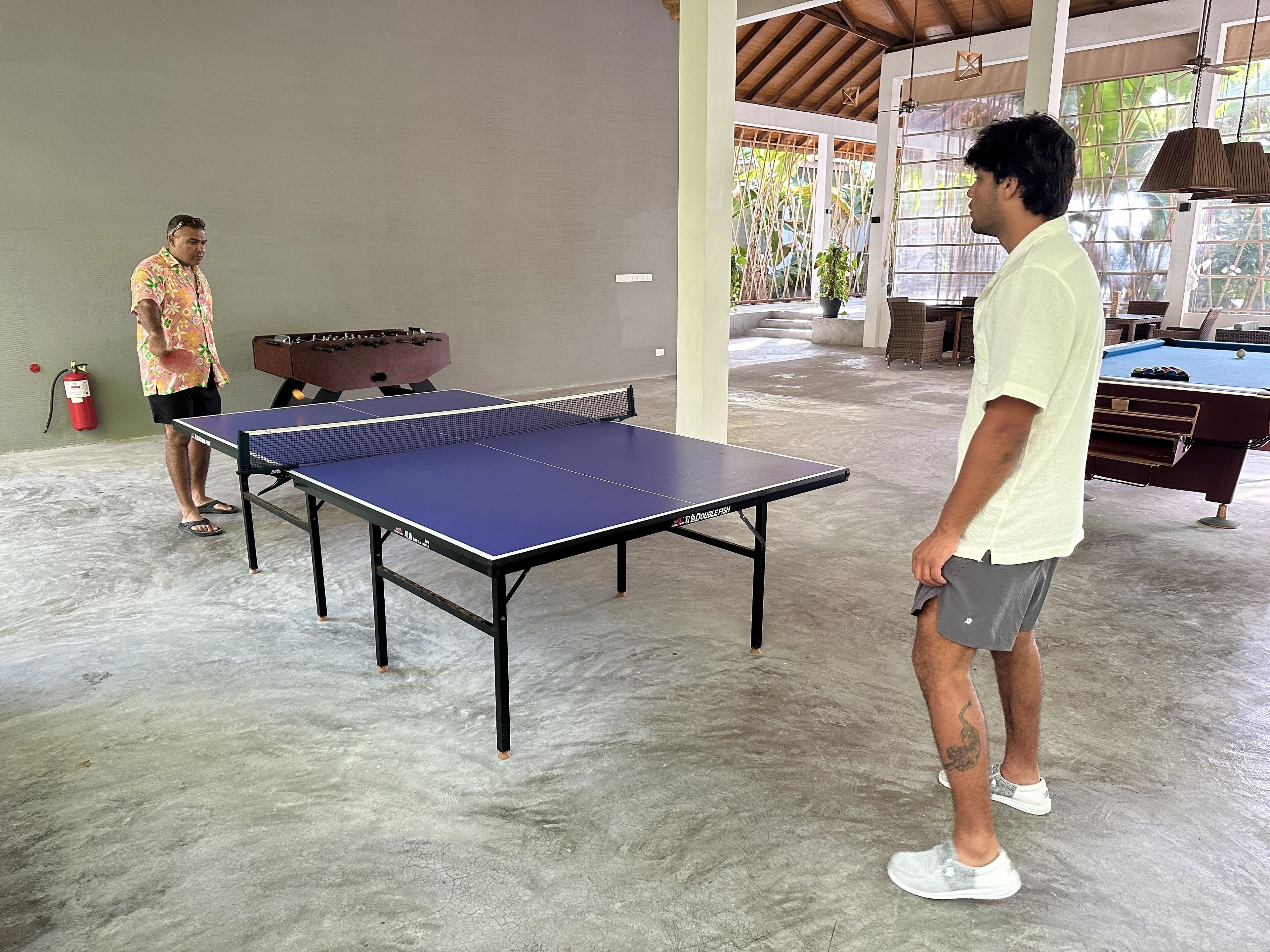 Una mesa de ping pong en un amplio espacio interior con piso de concreto, una mesa de futbolín y una mesa de billar.
