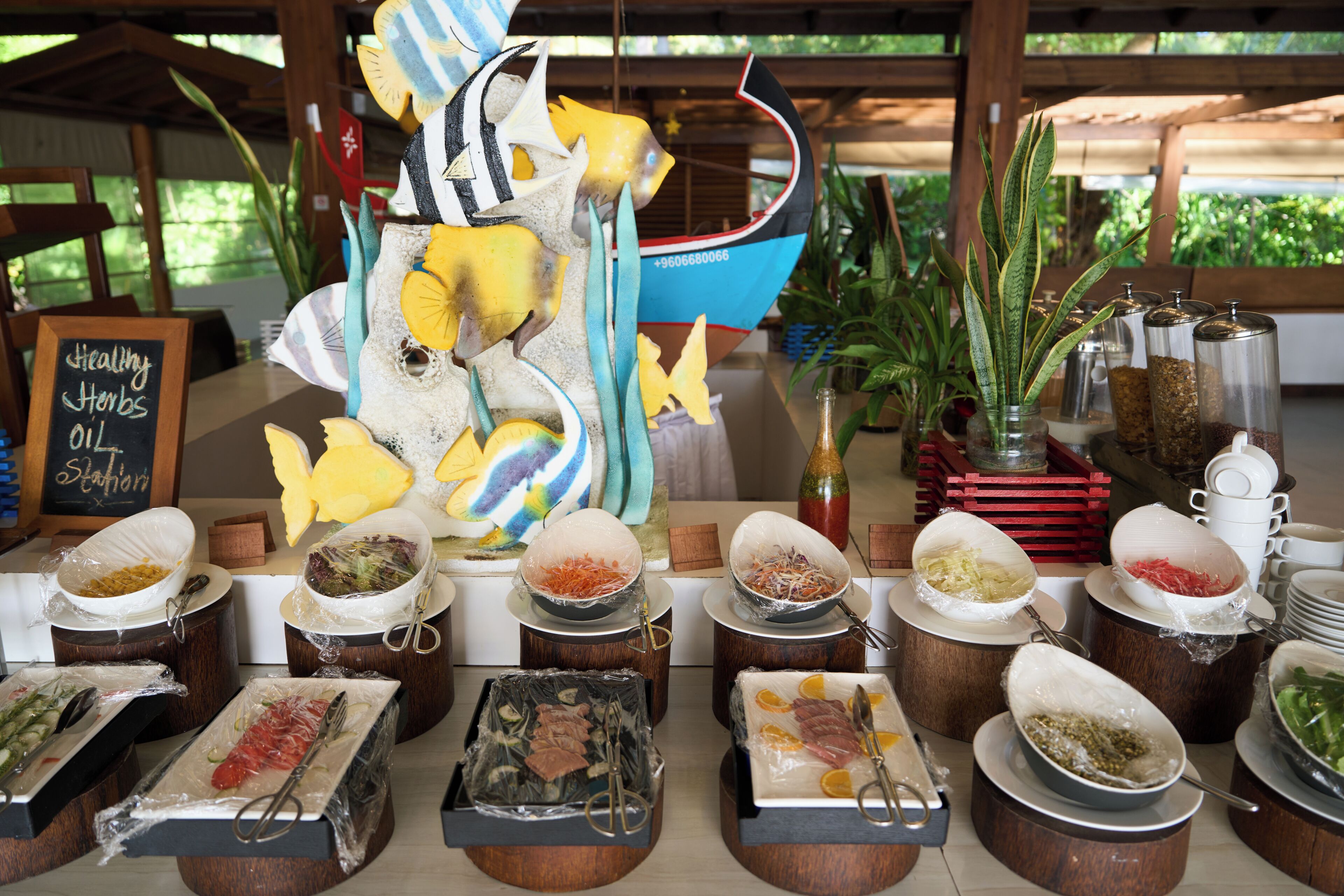 Disposição de buffet com diversos alimentos em recipientes fechados, um barco decorativo e uma placa indicando azeite de ervas saudáveis.