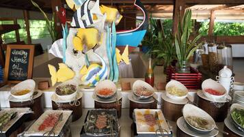 Disposição de buffet com diversos alimentos em recipientes fechados, um barco decorativo e uma placa indicando azeite de ervas saudáveis.