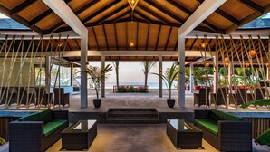 An outdoor covered seating area with wicker furniture, a view of the ocean, and a wooden ceiling structure. - Fiyavalhu Resort Maldives (Mandhoo)