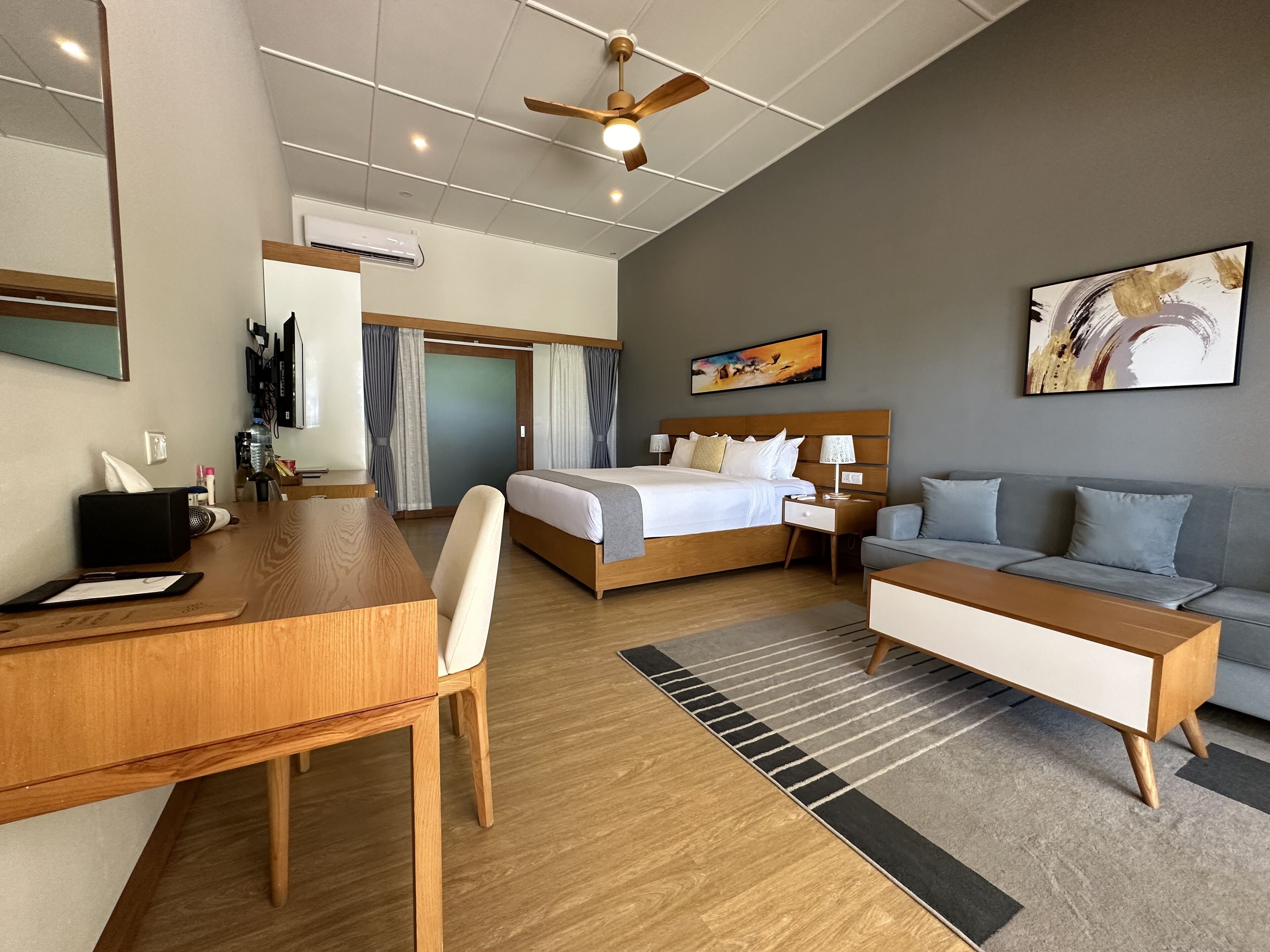 A hotel room with a bed, a desk, a sofa, a ceiling fan, and a painting on the wall.