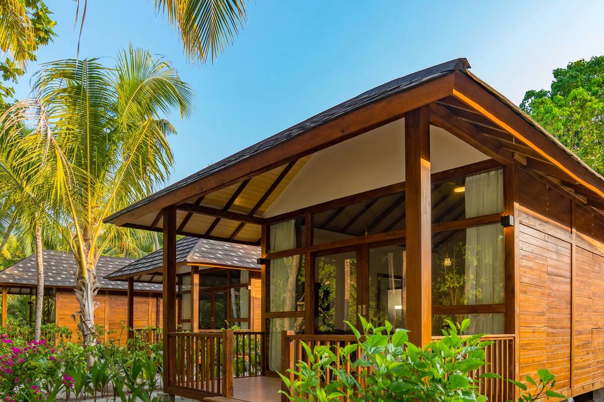 A wooden bungalow with a porch, large windows, and a view of palm trees and greenery.