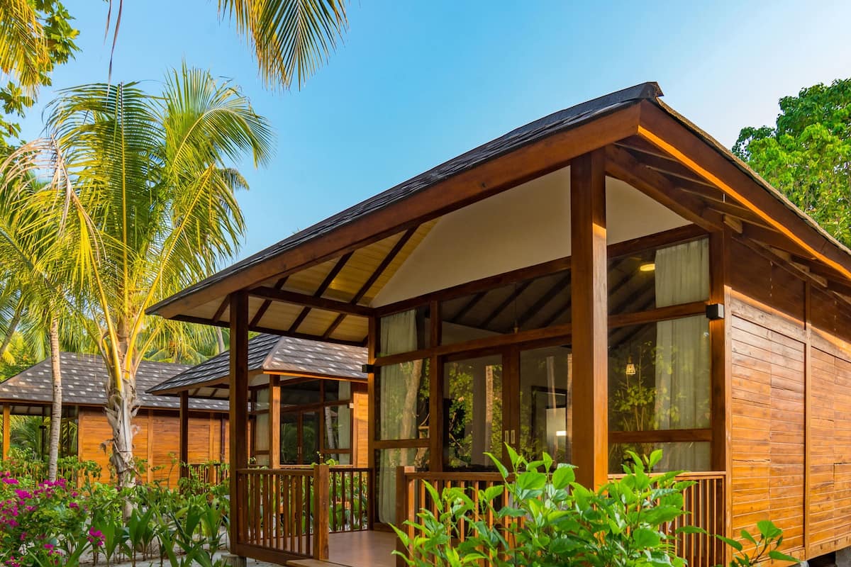Un bungalow en bois doté d’une véranda, de grandes fenêtres et offrant une vue sur des palmiers et une végétation luxuriante.