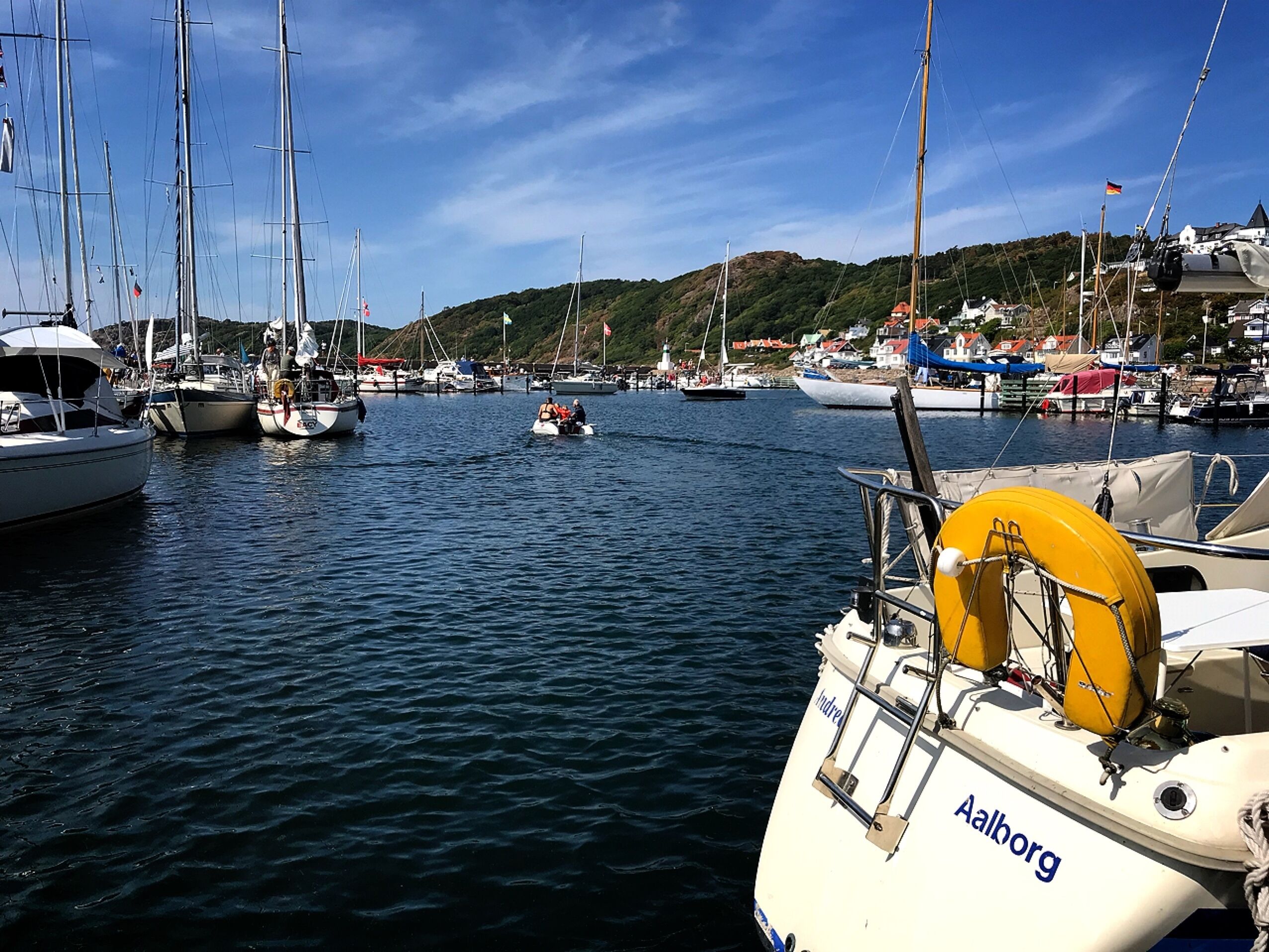 A marina with multiple sailboats docked, a small motorboat in the foreground, and a hill with houses in the background.