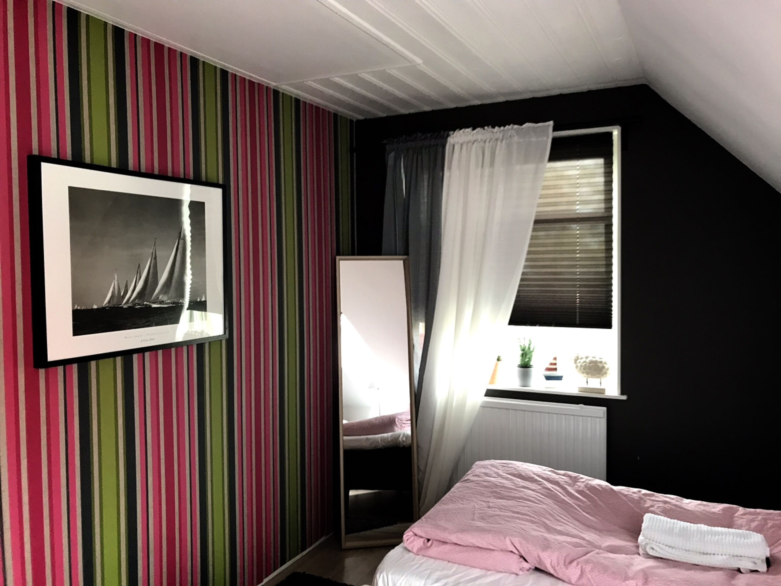 A bedroom with striped wallpaper, a bed with pink bedding, a window with curtains, and a framed picture on the wall.