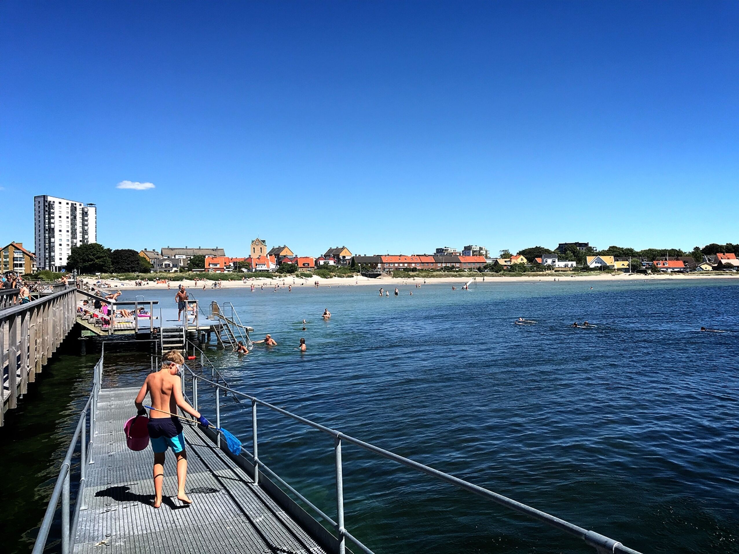 Une jetée s’avançant dans une mer calme, avec des personnes qui nagent et prennent des bains de soleil sur la plage en arrière-plan.