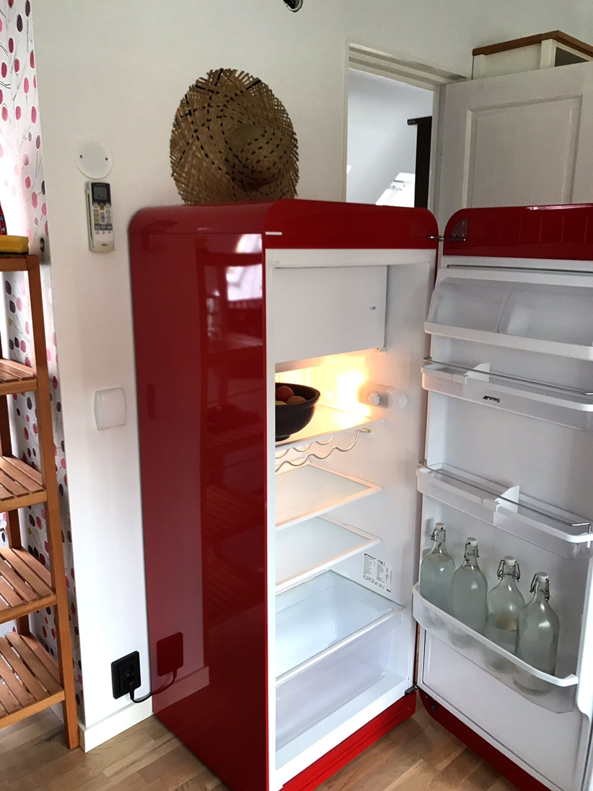 A red refrigerator with a basket on top, an open door revealing shelves and bottles, and a wooden shelf unit to the left.