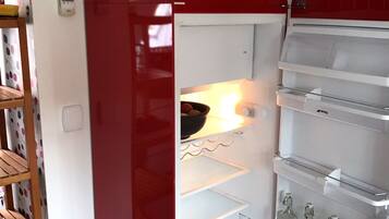 A red refrigerator with a basket on top, an open door revealing shelves and bottles, and a wooden shelf unit to the left.