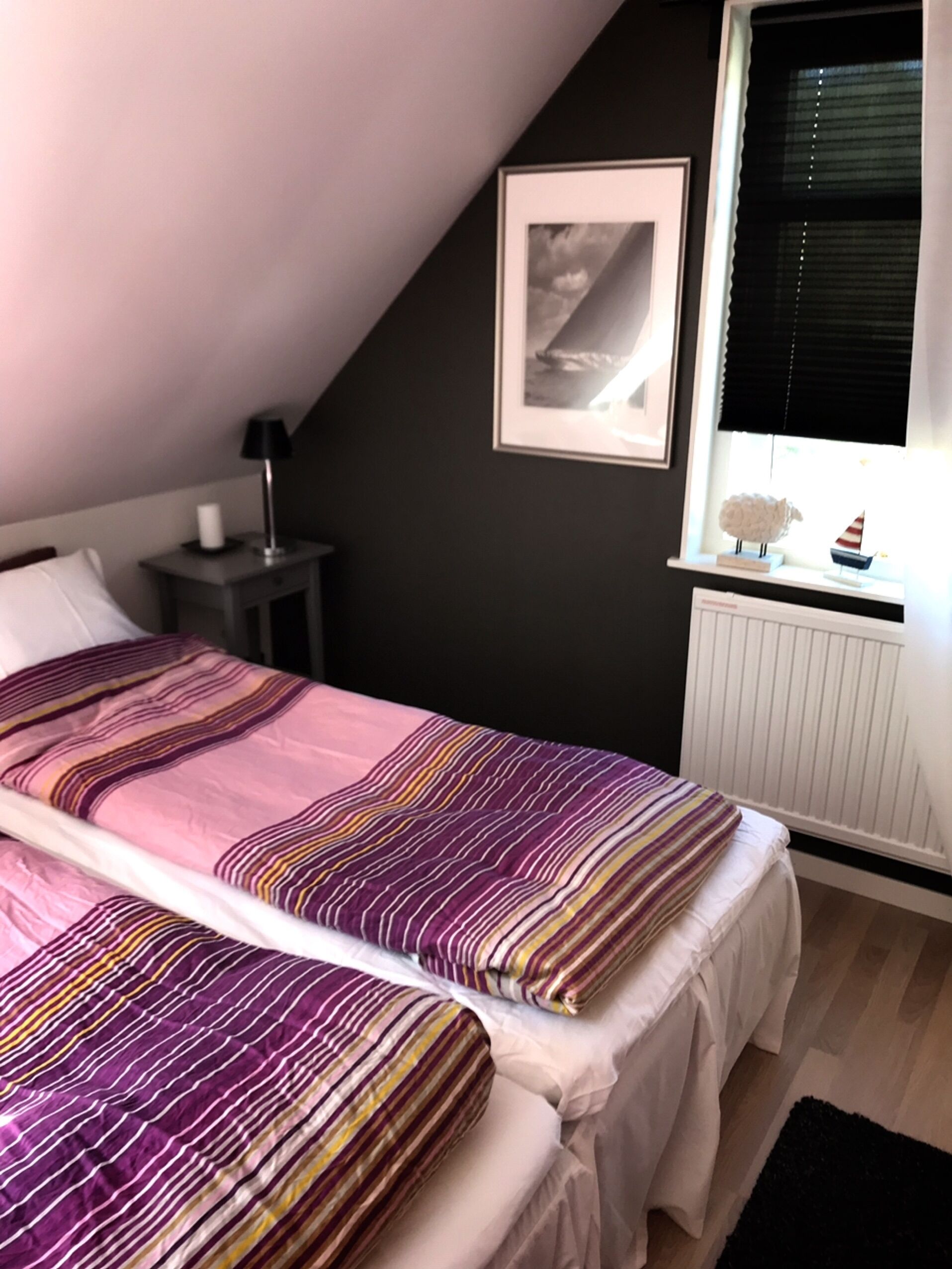 A bedroom with a sloped ceiling, a bed with striped bedding, a bedside table with a lamp, a framed picture on the wall, and a window with blinds.