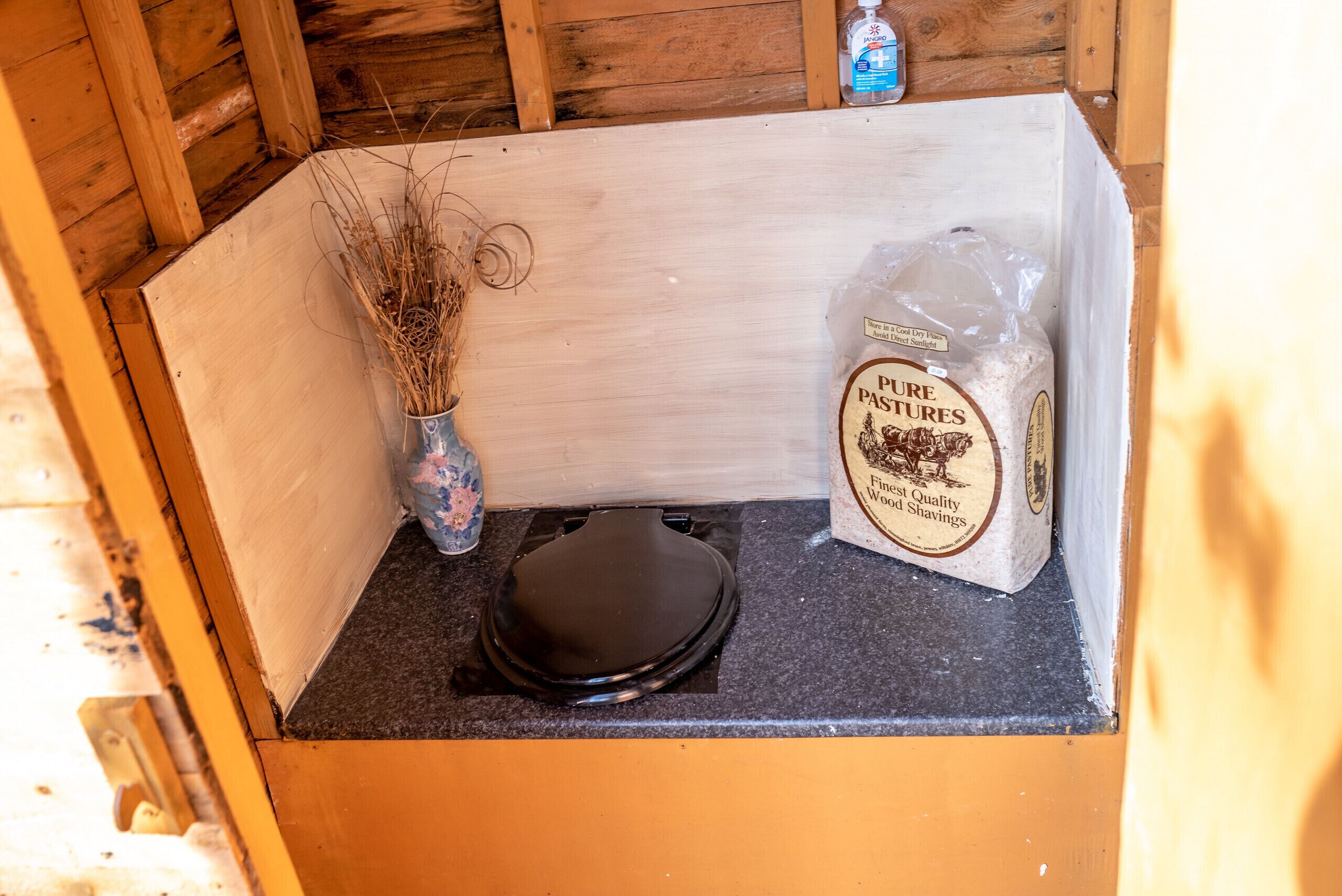 Een houten kastje met een zwart deksel, een vaas met gedroogde planten, en een zak Pure Pastures Fine Quality Wood Shavings.