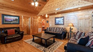 Interior acogedor de una cabaña de madera con techo de madera, ventilador de techo, chimenea, comedor y sala de estar con dos sofás y una mesa de centro.