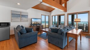 Un'ampia zona giorno con soffitto alto, travi in legno, una grande finestra con vista sull'oceano, una televisione e un angolo salotto.