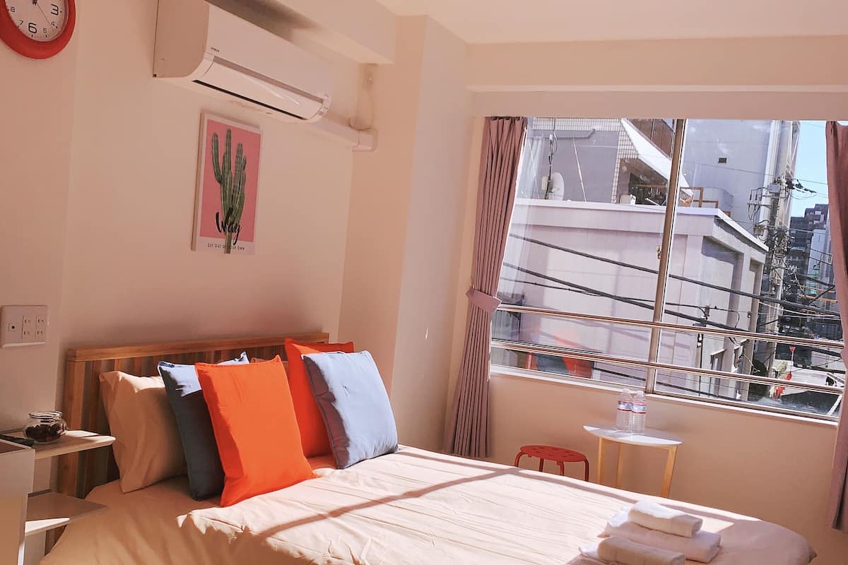 Habitación de hotel con cama, almohadas, un reloj, aire acondicionado, una ventana con cortinas y una mesita con botellas.