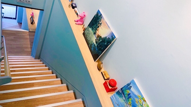 Un escalier avec des marches en bois et une rampe, menant à un palier avec un mur bleu et un panneau indiquant : « ATTENTION, DANGER AU-DESSUS ».