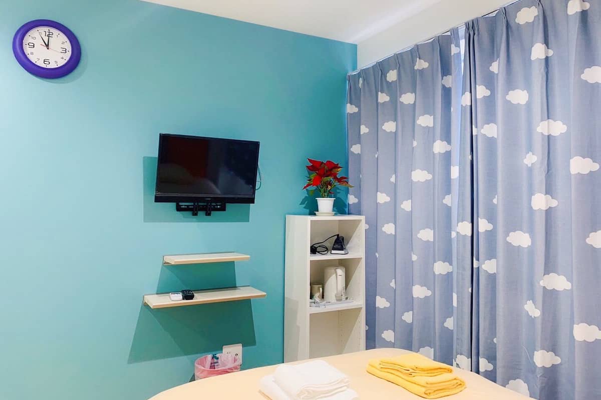 Una habitación con paredes azul verdosas, un televisor montado, una estantería con una planta en maceta y una cama con toallas dobladas.