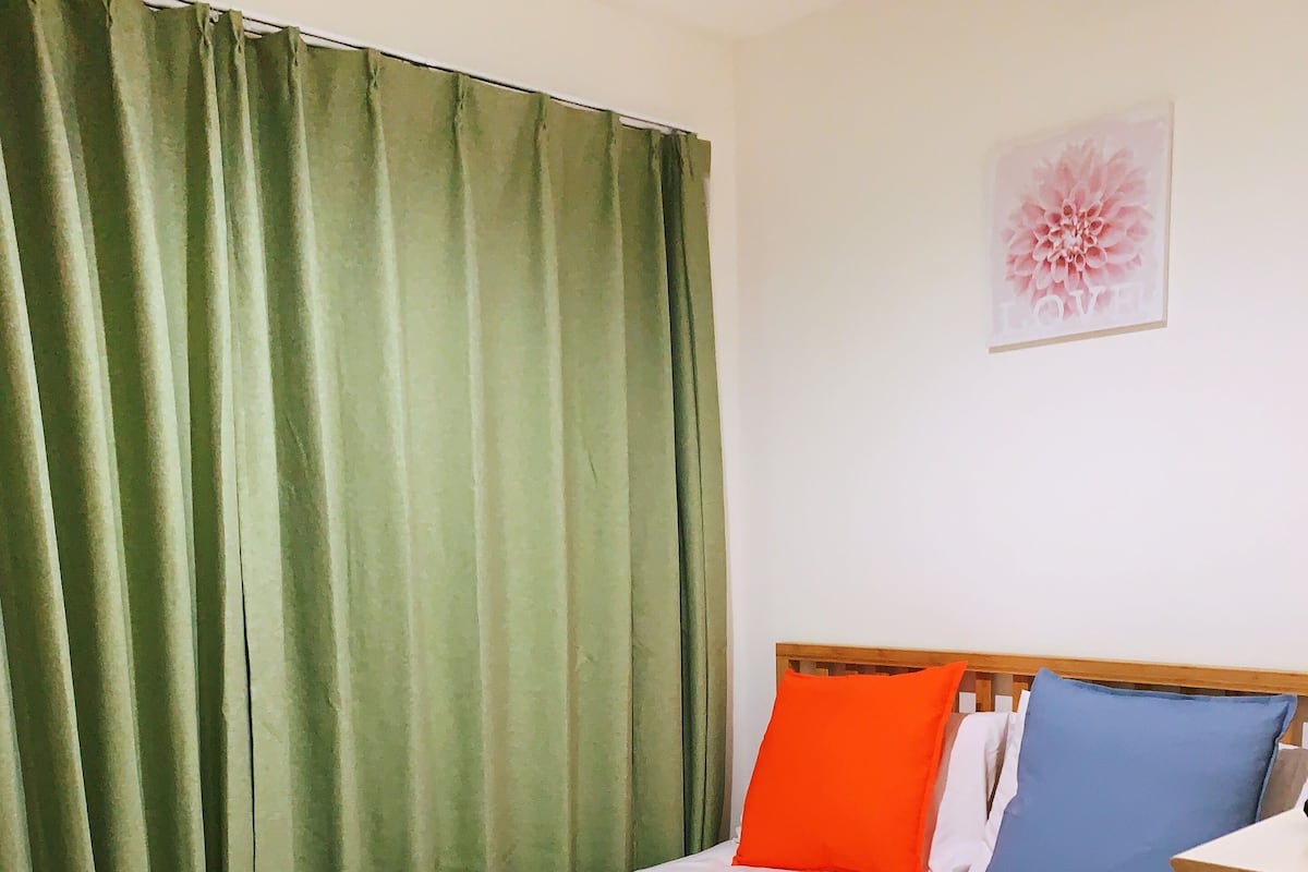 Una cama bien tendida con sábanas blancas, una cortina verde y un cuadro floral en la pared.