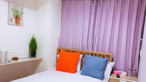 Un lit bien fait, avec du linge de lit blanc, une tête de lit en bois et deux oreillers (un orange, un bleu). On aperçoit une petite table de chevet avec une télécommande, une bouteille d’eau et une boîte. Une plante en pot est posée sur le mur, à gauche du lit.