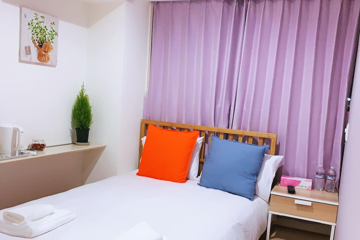 Una cama bien hecha con sábanas blancas, cabecera de madera y dos almohadas (una naranja y una azul). Se ve una mesita de noche con un control remoto, una botella de agua y una caja. Hay una planta en maceta en la pared a la izquierda de la cama.