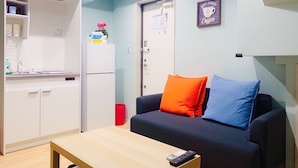 Un espace de vie compact comprenant un coin cuisine, un canapé avec deux coussins, une petite table et un tableau encadré accroché au mur.