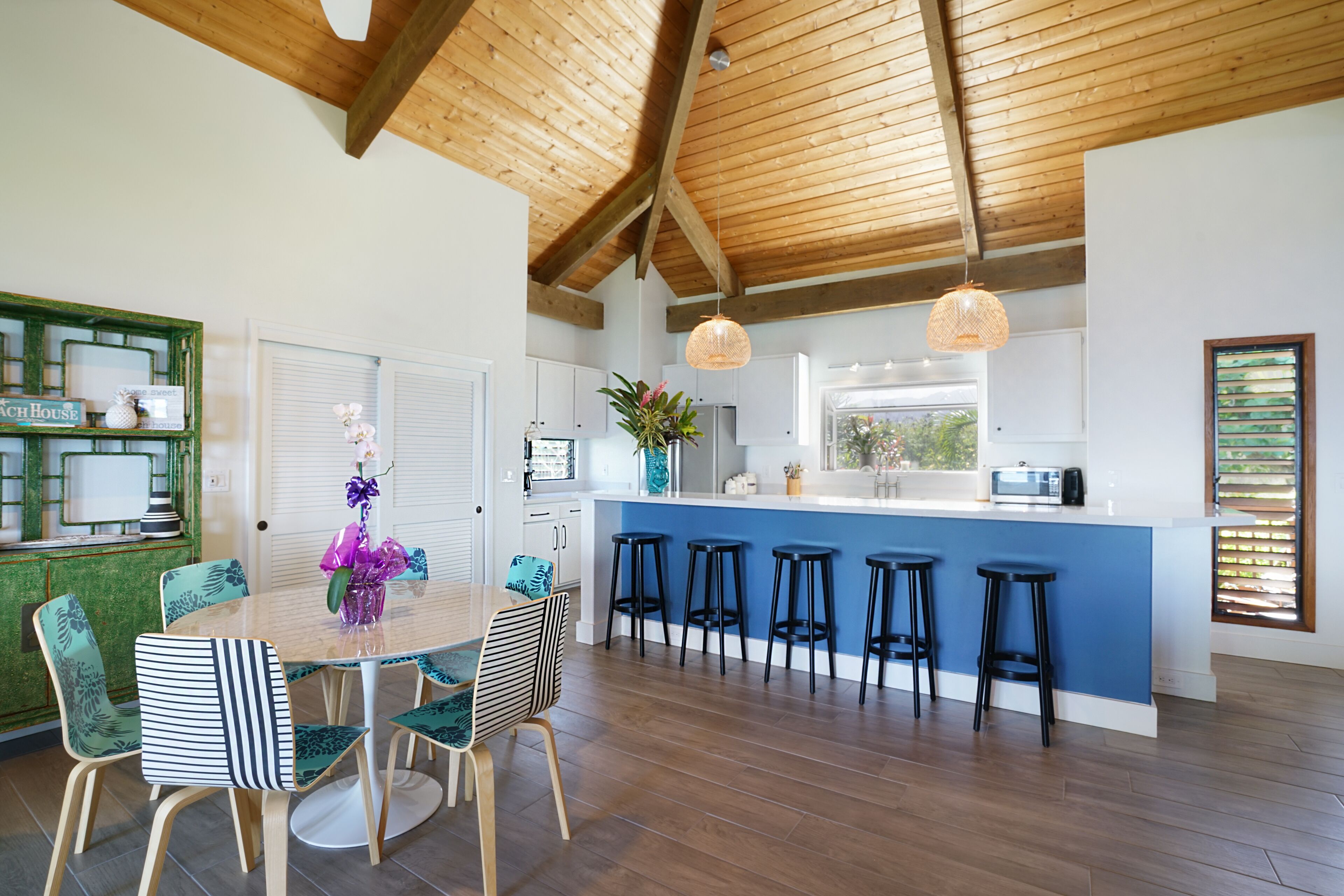 A modern kitchen with a blue island, wooden ceiling beams, and a dining area with striped chairs.
