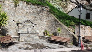 Zona con panchine in pietra, uno sfondo a muro in pietra, una piccola costruzione in pietra e un faro.