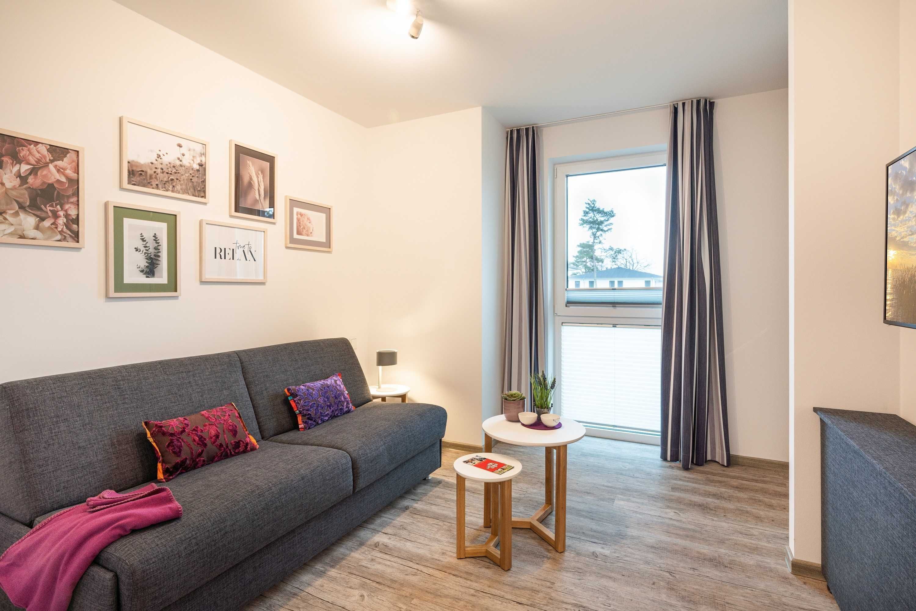 Un salon moderne avec un canapé gris, une petite table ronde ornée d’une plante et des tableaux encadrés accrochés au mur.