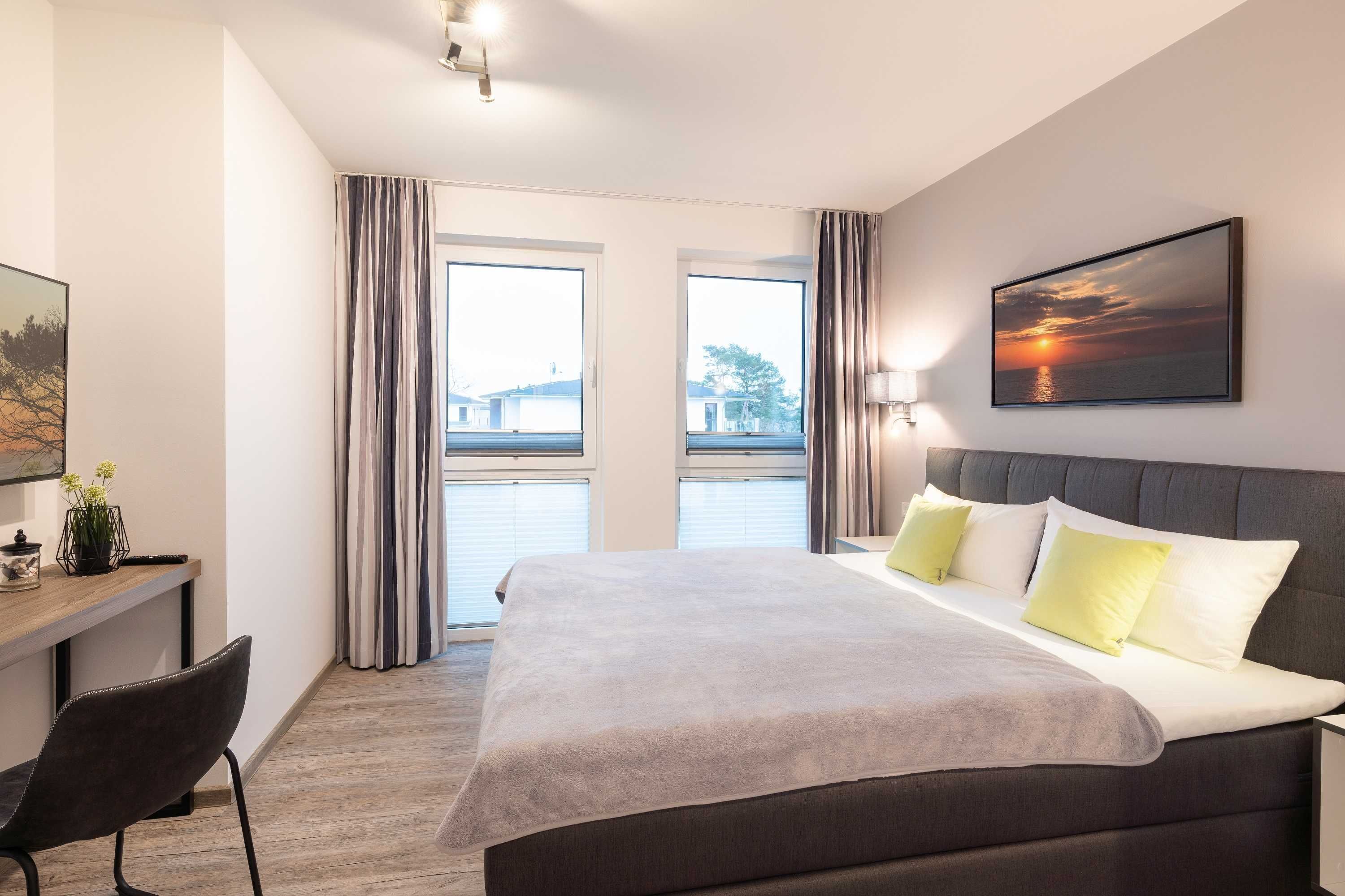 Quarto de hotel moderno com uma cama grande, uma escrivaninha e vista para o mar pelas janelas.
