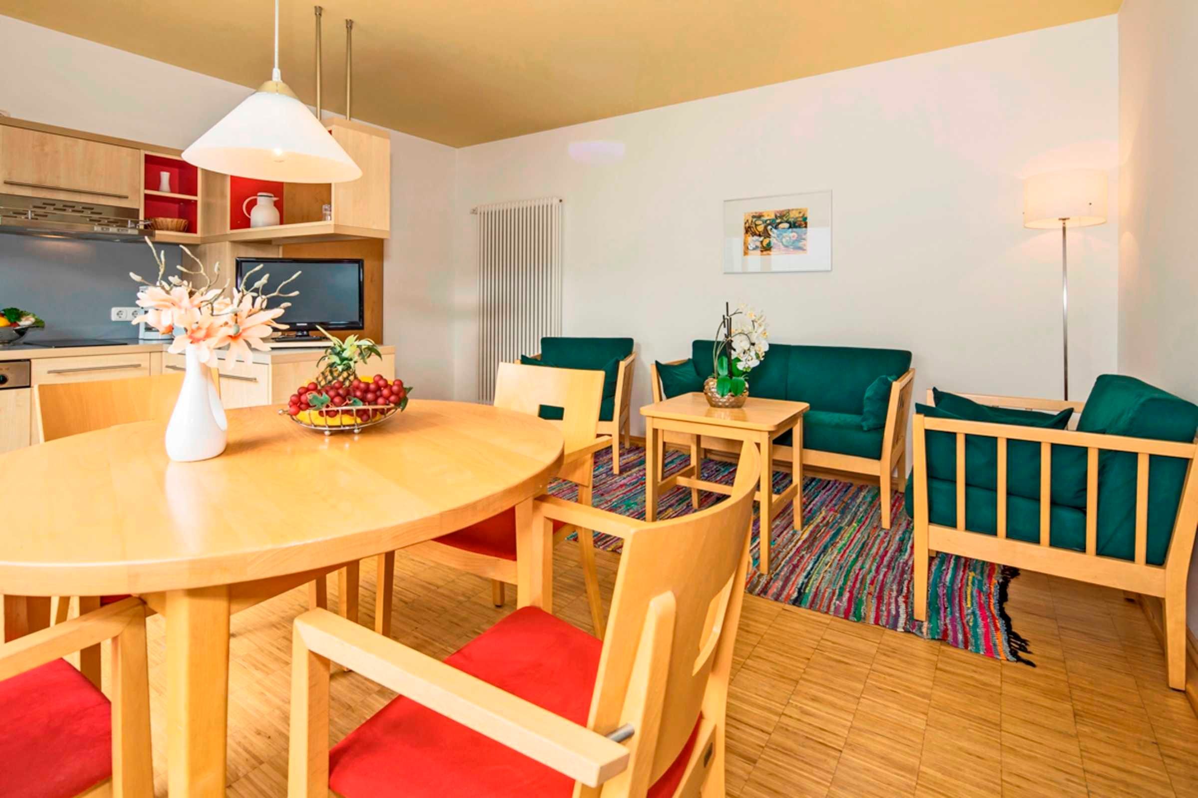 Una sala moderna con una mesa de comedor de madera, sillas con cojines rojos, un sofá verde y una pequeña mesita de noche con un florero.