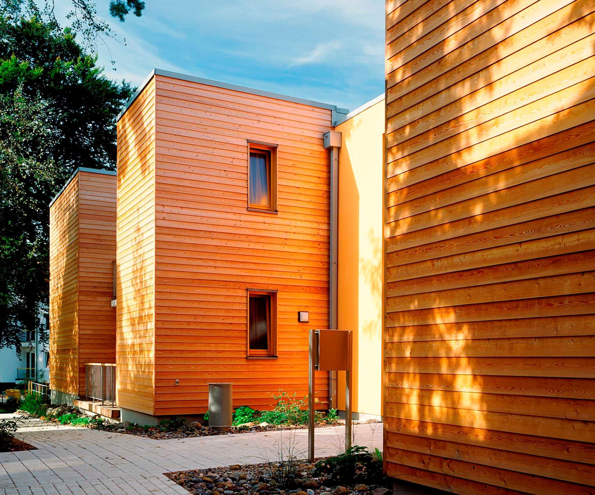 Un bâtiment moderne doté d’un revêtement extérieur en bois disposé horizontalement, de deux fenêtres apparentes et d’un petit espace extérieur agrémenté d’un chemin et de quelques plantes.