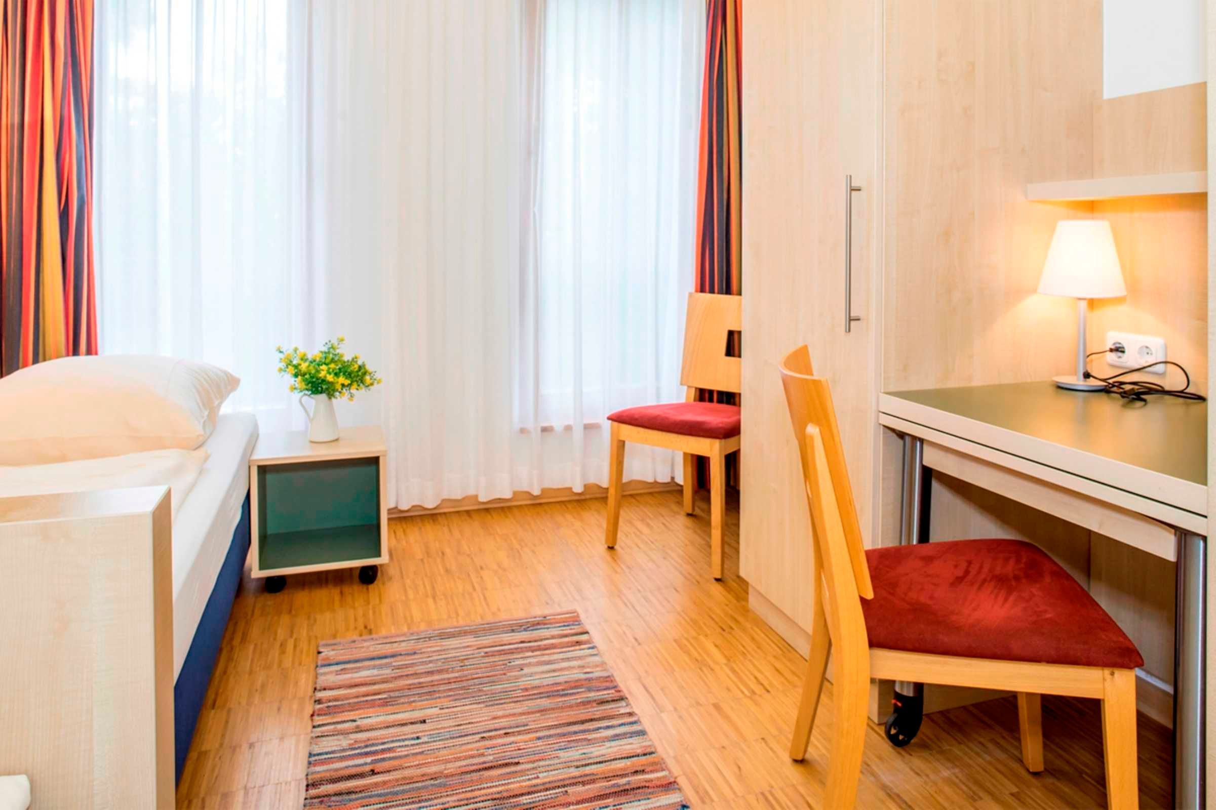 Quarto de hotel com cama, escrivaninha, cadeira, mesinha de apoio com abajur e janela com cortinas.