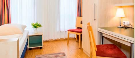 Quarto de hotel com cama, escrivaninha, cadeira, mesinha de apoio com abajur e janela com cortinas.