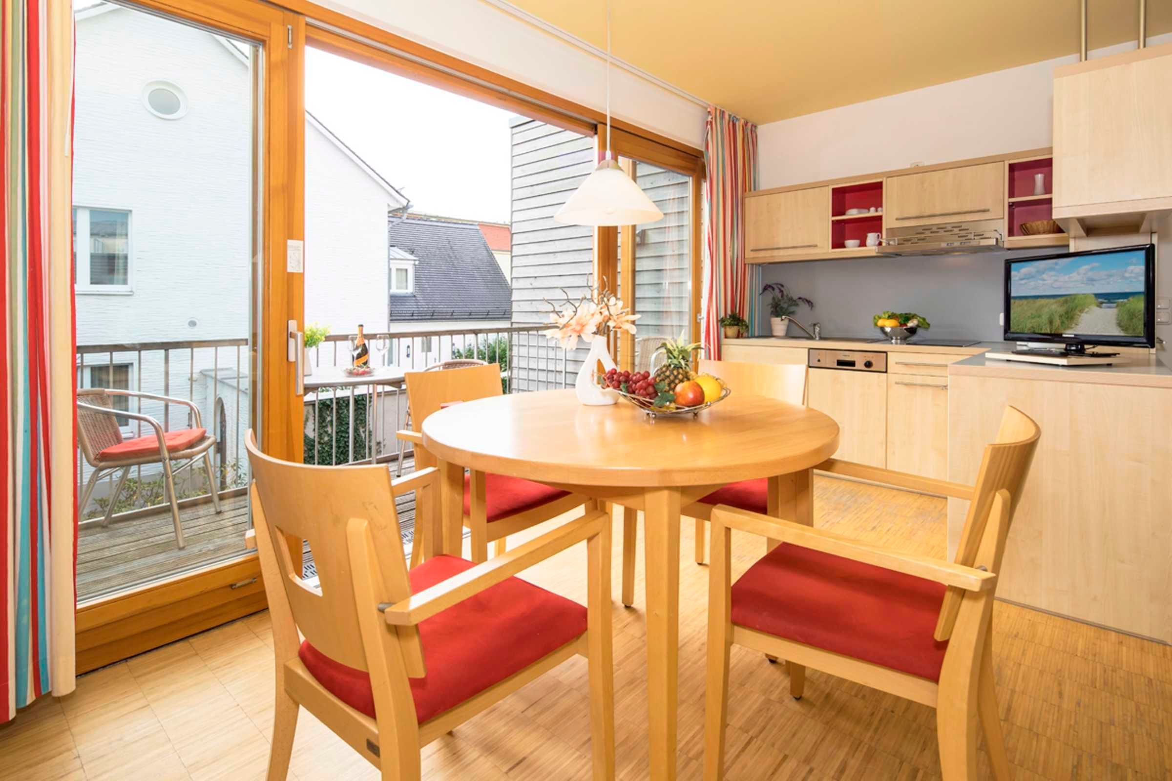 Un coin repas avec une table ronde, des chaises en bois avec des coussins rouges, un panier de fruits et un téléviseur posé sur un meuble.