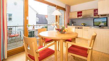 Un coin repas avec une table ronde, des chaises en bois avec des coussins rouges, un panier de fruits et un téléviseur posé sur un meuble.