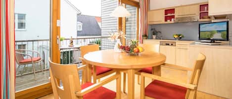 A dining area with a round table, wooden chairs with red cushions, a fruit bowl, and a television on a cabinet.