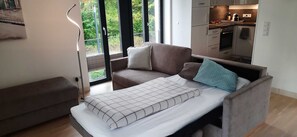 Ein modernes Schlafzimmer mit einem Bett, einem Sofa und Blick auf eine Küche sowie Grünflächen im Freien.