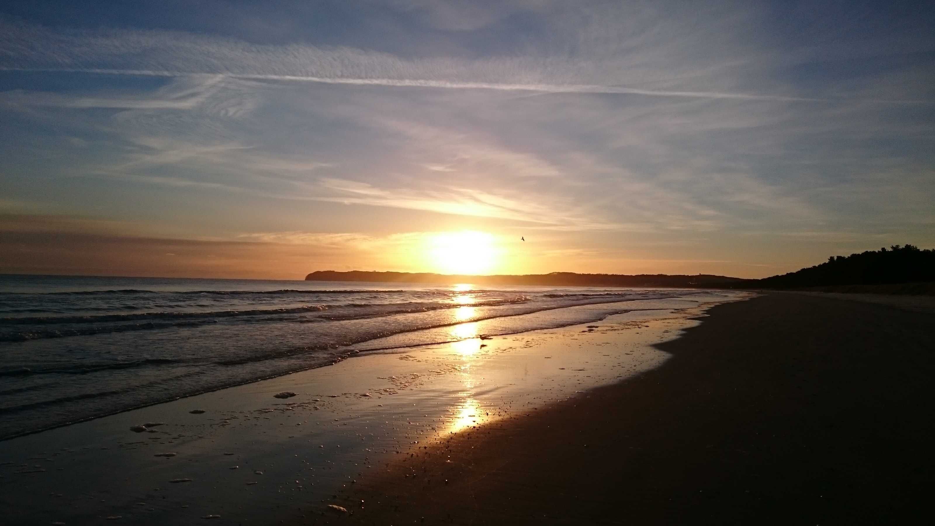 Ein Strand mit untergehendem Sonnenuntergang am Horizont, ruhigen Wellen und wolkenlosem Himmel.