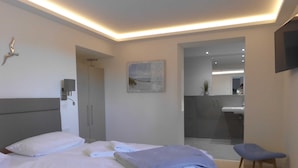 Habitación de hotel moderna con cama, una silla, un escritorio y un baño visible a través de una puerta de vidrio.