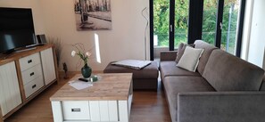 Ein modernes Wohnzimmer mit einem Flachbildfernseher, einem Holztisch, einer Couch und einem großen Fenster mit Blick auf Grünflächen.