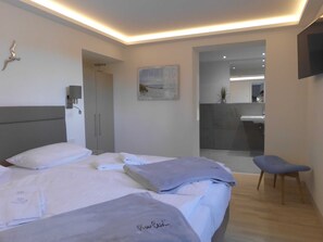 Ein modernes Hotelzimmer mit Bett, Sessel, Schreibtisch und einem durch eine Glastür einsehbaren Badezimmer.