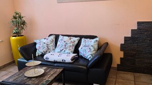 A living room with a black leather sofa, floral cushions, a small coffee table, a yellow plant, and a painting of a beach scene on the wall. - Kos - sparrow lust - sparrow lust (Koserow)